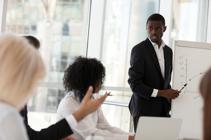 African American mentor or coach give flipchart presentation to colleagues, answer employees question, black businessman present business plan, explaining project or strategies to office workers